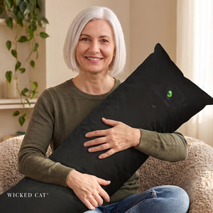A smiling woman with short white hair sits on a textured beige couch holding a long black throw pillow. The pillow features a dark cat design with glowing green eyes.