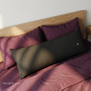 A dark olive green lumbar pillow with a small green cat face graphic, styled on a bed with burgundy sheets and matching pillows next to a wooden headboard. The brand name 'WICKED CAT' is visible in the bottom-left corner.