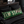 A rectangular green throw pillow with distressed cream lettering reading 'ELM STREET' and red blood-like drips, resting on a bed with black bedding in a dark bedroom setting. Wicked Cat branding appears at the bottom left.