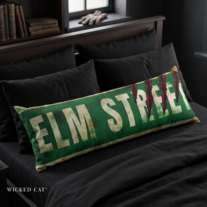 A rectangular green throw pillow with distressed cream lettering reading 'ELM STREET' and red blood-like drips, resting on a bed with black bedding in a dark bedroom setting. Wicked Cat branding appears at the bottom left.