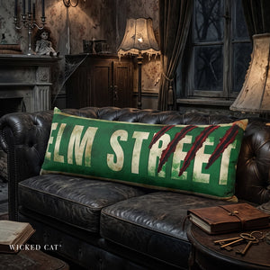 A green rectangular throw pillow with distressed white 'ELM STREET' lettering and red claw marks, resting on a dark leather sofa in a moody, vintage-style room.