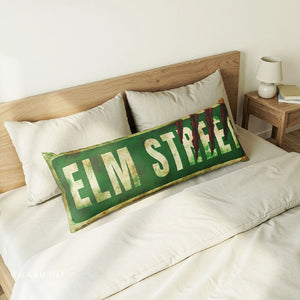 A rectangular decorative throw pillow styled on a bed, featuring a vintage green 'ELM STREET' sign design with distressed cream lettering and gold-toned edging. The pillow is shown on a bed with white linens and a wooden headboard.