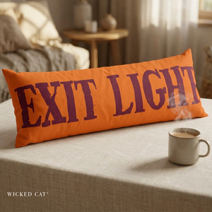 An orange rectangular throw pillow with the words 'EXIT LIGHT' printed in large distressed purple letters, resting on a beige tablecloth in a softly lit living room. A ceramic mug sits nearby, and the brand name 'WICKED CAT' appears in the lower left corner.