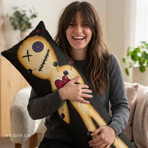 A woman smiling while holding a long black throw pillow with a stitched voodoo doll design in a cozy indoor setting. The pillow features a tan face and body with colorful button and pin details.