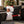 A square decorative throw pillow featuring a festive Christmas design, resting on a dark gray sofa in a decorated living room. The pillow displays a split design: one side shows a cartoon-style Black Santa Claus with a white beard and hat, and the other side is black with white snowflakes and a hand holding a champagne glass.