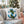 A square white throw pillow with a blue anchor and green holly design, styled on a light gray sofa in a cozy living room with a decorated Christmas tree in the background.