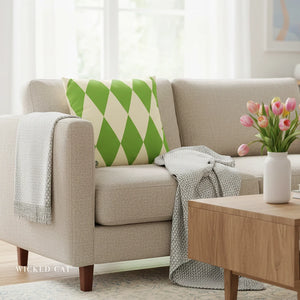 Beige sofa with a green and white patterned pillow, gray blanket, and wooden coffee table with flowers in a room.