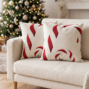 Two square throw pillows with a red and cream candy cane pattern, styled on a textured beige armchair in front of a decorated Christmas tree.