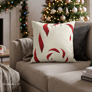A square throw pillow with a red abstract candy cane pattern on an off-white background, styled on a beige sofa in a cozy living room setting with a Christmas tree in the background. The brand name 'WICKED CAT' is visible in the bottom-left corner.