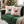 Two square cream throw pillows with a pink and green candy cane and holly design, resting on a green velvet sofa in a cozy, blurred living room setting with a Christmas tree in the background.