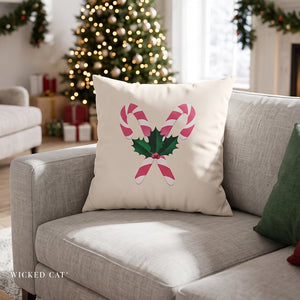 A square cream throw pillow with a pink and green candy cane and holly design, resting on a light gray sofa in a decorated living room with a Christmas tree in the background.