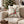 A square throw pillow with a red and green abstract holiday pattern on an off-white background, styled on a beige sofa in front of a blurred Christmas tree.