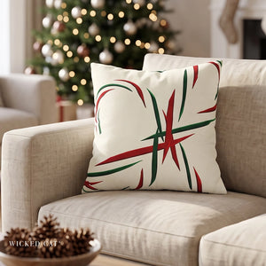 A square throw pillow with a red and green abstract holiday pattern on an off-white background, styled on a beige sofa in front of a blurred Christmas tree.