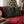 Two black Christmas throw pillows with green holly and red berries printed designs, resting on a red velvet sofa in a living room with a blurred decorated Christmas tree in the background. The brand name 'Wicked Cat' is visible in the bottom left corner.