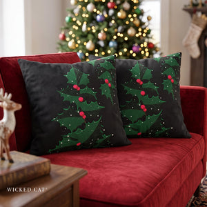 Two black Christmas throw pillows with green holly and red berries printed designs, resting on a red velvet sofa in a living room with a blurred decorated Christmas tree in the background. The brand name 'Wicked Cat' is visible in the bottom left corner.