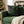 A square black throw pillow with a green holly and red berries pattern, styled on a green velvet armchair in a cozy, warm Christmas living room setting.
