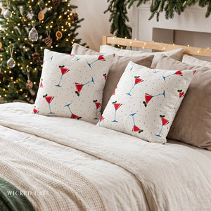 Two square decorative throw pillows with a white background and a red, blue, and green martini glass pattern, styled on a bed with beige sheets and pillows in a cozy holiday bedroom setting with a decorated Christmas tree in the background.