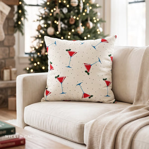 A square throw pillow with a holiday martini pattern, resting on a beige sofa in a cozy living room with a blurred Christmas tree in the background.