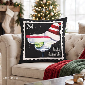 A square decorative throw pillow with a black background and scalloped white border, featuring an illustration of a margarita glass with a red rim, lime slice, Santa hat, and the text '25¢ Merry Margarita.' The pillow is styled on a beige tufted sofa in a cozy living room with a Christmas tree and stockings in the background.