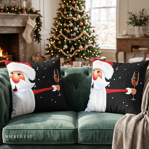 Two decorative throw pillows featuring a festive Christmas design of Santa Claus holding a glass of champagne on a black background with white snowflakes, displayed on a green velvet sofa in a decorated living room with a fireplace and a Christmas tree.