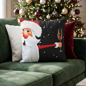 A decorative throw pillow featuring a festive Christmas design of Santa Claus holding a champagne glass, styled on a green velvet sofa in front of a lit Christmas tree.