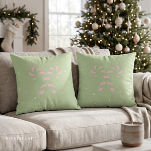 Two sage green throw pillows with a pink Christmas tree design, displayed on a beige sofa in a cozy, decorated living room with a Christmas tree in the background.