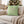 A sage green throw pillow with a pink reindeer face design and white snowflakes, resting on a beige sofa in a cozy living room with a Christmas tree in the background.