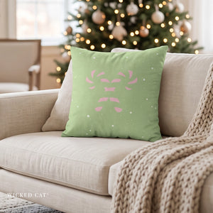 A sage green throw pillow with a pink reindeer face design and white snowflakes, resting on a beige sofa in a cozy living room with a Christmas tree in the background.