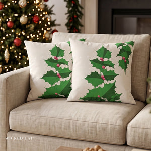 Two square white throw pillows with a green holly and red berries pattern, styled on a beige fabric sofa in a cozy living room setting with a decorated Christmas tree in the background.