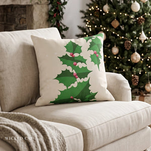 A square decorative throw pillow with a green holly and white cloud-like border design, resting on a light beige sofa in a cozy living room setting with a Christmas tree in the background. The brand name 'WICKED CAT' is visible in the bottom left corner.