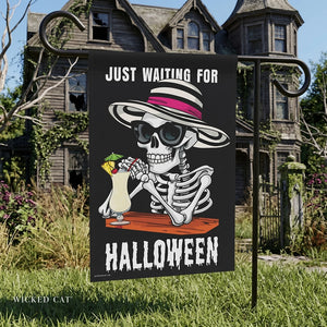 Yard flag with a skeleton wearing a hat and sunglasses drinking a pina colada and text that reads just waiting for summer, in front of a haunted house