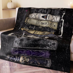A black throw blanket draped over a sofa, printed with illustrated witch books and spells including 'The Glass Cauldron', 'Exorcism', 'Wicked Curses', and 'Wicked Cat'.