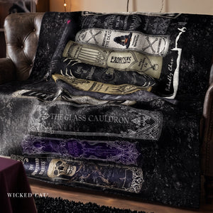 A dark throw blanket draped over a brown leather armchair, printed with illustrated potion bottles and spells from Wicked: The Great Dictation, including The Glass Cauldron, Horcruxes, and The Wizard's Chamber. The blanket has a distressed black and gray texture with white and purple accents.