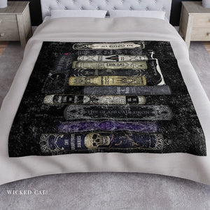 A bed dressed in white bedding with a large printed blanket or comforter featuring stacked gothic-style book spines in black, beige, gray, and purple. The room includes light wood nightstands and a gray rug.