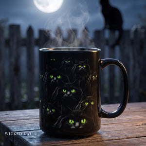 A black ceramic mug with a handle, decorated with multiple illustrated black cats with glowing green eyes, sitting on a wooden table with steam rising. A blurred nighttime fence, full moon, and a cat silhouette are in the background.