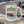 A white ceramic mug with a colorful shield-shaped graphic reading 'Instructor Toonce's Stunt Driving' and showing a pink race car, placed on a concrete barrier with a blurred racetrack and race cars in the background.