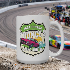 A white ceramic mug with a colorful shield-shaped graphic reading 'Instructor Toonce's Stunt Driving' and showing a pink race car, placed on a concrete barrier with a blurred racetrack and race cars in the background.