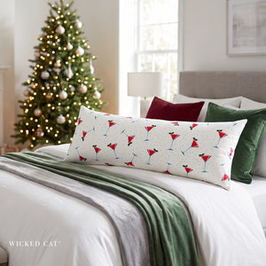 A long rectangular throw pillow with a white background and a red, green, and martini cocktail print, styled on a bed in a cozy bedroom with a decorated Christmas tree in the background.