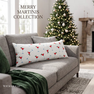 A rectangular lumbar throw pillow with a festive holiday print of red and blue martinis on a white background, styled on a gray sofa in a cozy living room with a Christmas tree in the background. The brand 'Wicked Cat' is visible in the bottom left corner.