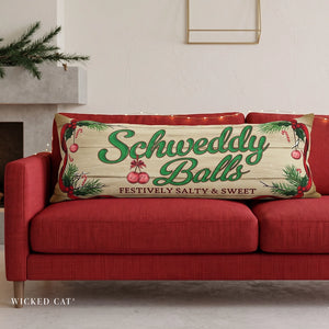 A rectangular decorative throw pillow with a rustic wood-grain background and the words 'Schweddy Balls' in green script, styled on a red sofa in a living room setting.