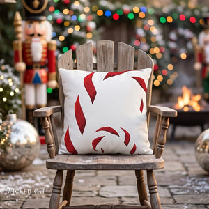A square white throw pillow with a red candy cane pattern, displayed on a rustic wooden rocking chair in a festive outdoor holiday setting with a fire pit, nutcracker, and Christmas lights.