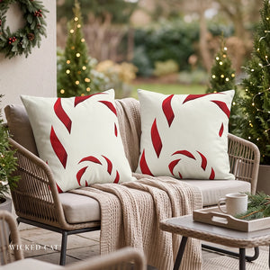 Two white outdoor throw pillows with a red candy cane pattern, styled on a wicker patio chair next to a beige knit blanket, with a festive garden background.