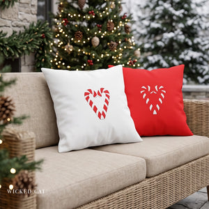 Two square throw pillows on a wicker sofa: one white with a red candy cane heart motif and one red with a white candy cane heart motif, set outdoors in front of a decorated Christmas tree.