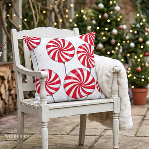 A square throw pillow with a red and white peppermint candy cane pattern sits on a distressed white wooden chair in an outdoor patio setting with a white faux fur throw blanket draped over the arm and a blurred Christmas tree in the background.