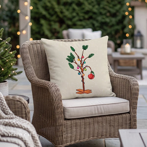 A square decorative throw pillow with a whimsical Christmas tree design, resting on a wicker patio chair in a cozy, softly lit outdoor setting.
