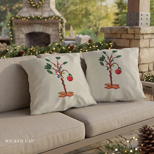 Two square throw pillows on a beige outdoor sofa, featuring a whimsical Christmas tree design with colorful ornaments and red apples. The background shows a cozy patio setting with a stone fireplace, green garland, and warm string lights.