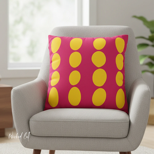 Pink pillow with yellow circles on a gray armchair in a room with a plant and window.