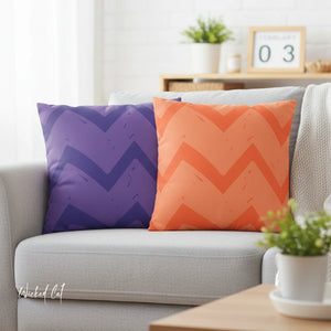 Two chevron patterned pillows, one purple and one orange, on a gray sofa with a blurred background.