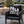 A black outdoor throw pillow with 'FALA LA LA LA LA LA' printed in bold white letters, styled on a dark wicker patio chair in front of a lit fire pit and Christmas tree.