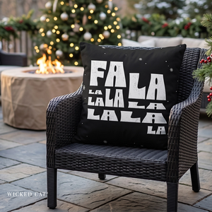 A black outdoor throw pillow with 'FALA LA LA LA LA LA' printed in bold white letters, styled on a dark wicker patio chair in front of a lit fire pit and Christmas tree.